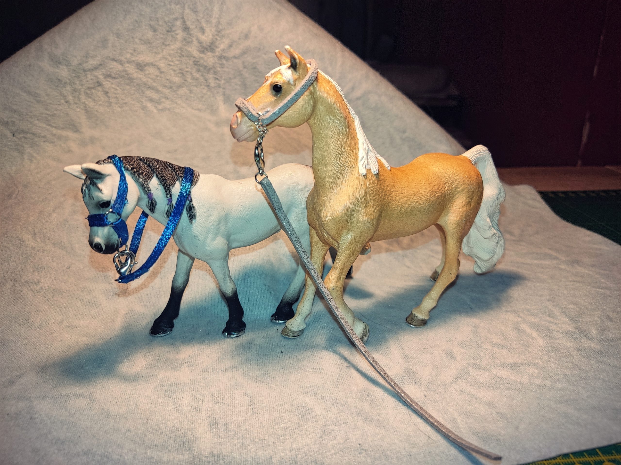 2 Schleichpferde stehen hintereinander, hinten ein weißes mit blauem Halfter und Zügeln, vorne ein hellbraunes mit hellbraun glitzerndem Halfter und Strick