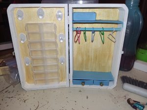 Eine aufrecht stehende kleine geöffnete Kiste. Darin links kleine Döschen als Schrank, rechts eine Kleiderstange, Minikleiderbügel, Ablagefächer und 3 Schubladen